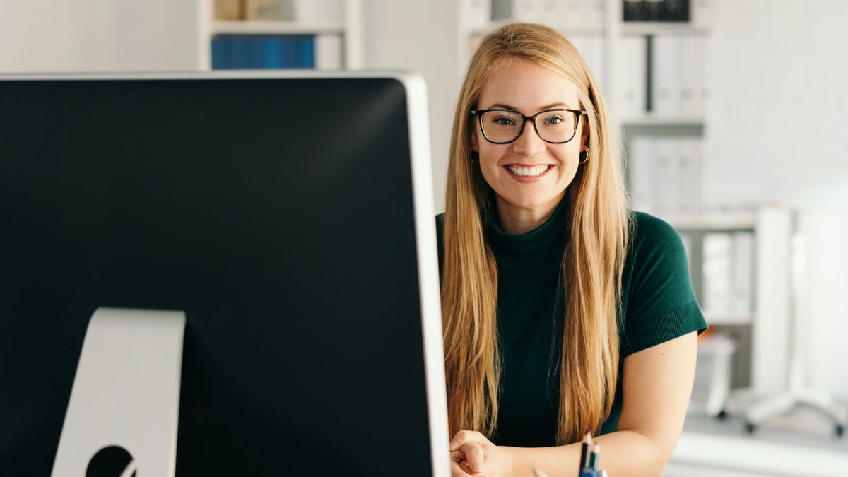 Friendly young secretary or personal assistant sitting working at a desktop computer in the office smiling at the camera